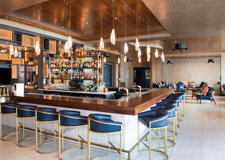 A modern bar with a wooden countertop, blue barstools, hanging lights, and a well-stocked array of bottles, situated in a spacious lounge area.