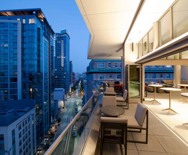 The image shows a modern cityscape at dusk with tall buildings and a stylish balcony or rooftop lounge area with seating and tables.