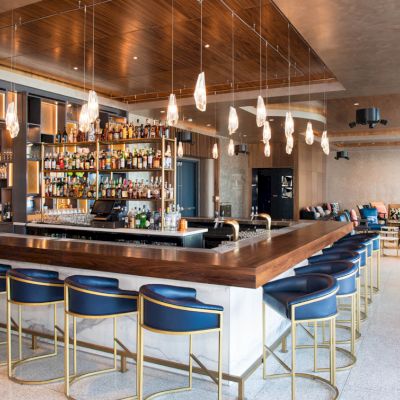 The image shows a modern bar with a wooden counter, blue and gold bar stools, hanging lights, and a fully stocked shelf of beverages in the background.