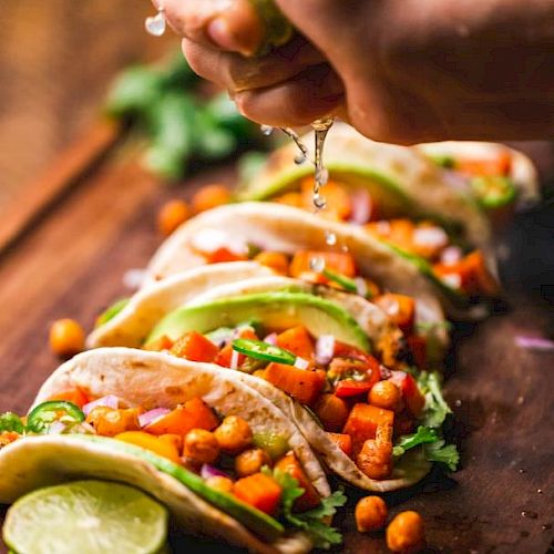The image shows a close-up of a hand squeezing lime juice over a row of vegetable tacos garnished with chickpeas, cilantro, and avocado slices on a wooden board.