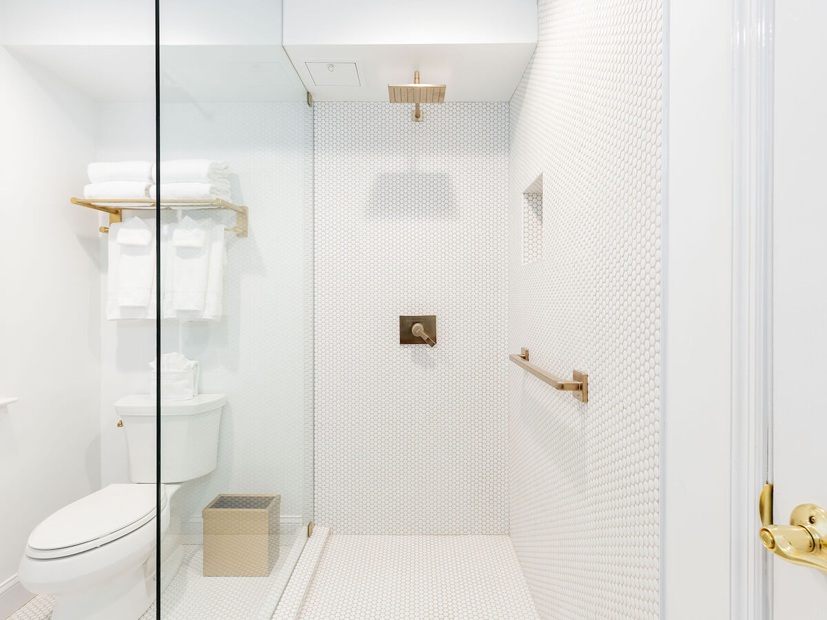 A modern bathroom with a glass shower, white walls, a toilet, and towels on a rack. The shower has a rainfall showerhead and gold fixtures.