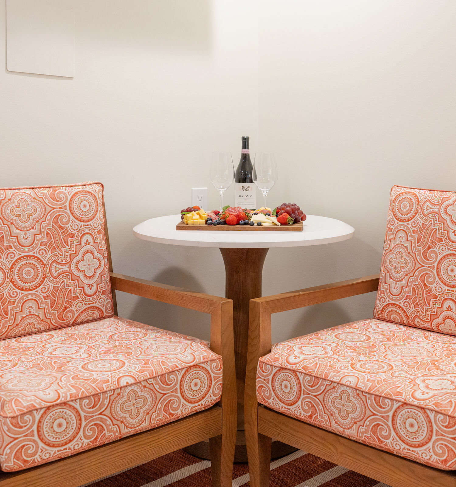Two orange patterned armchairs and a small round table with wine, glasses, and fruit are placed in a corner.
