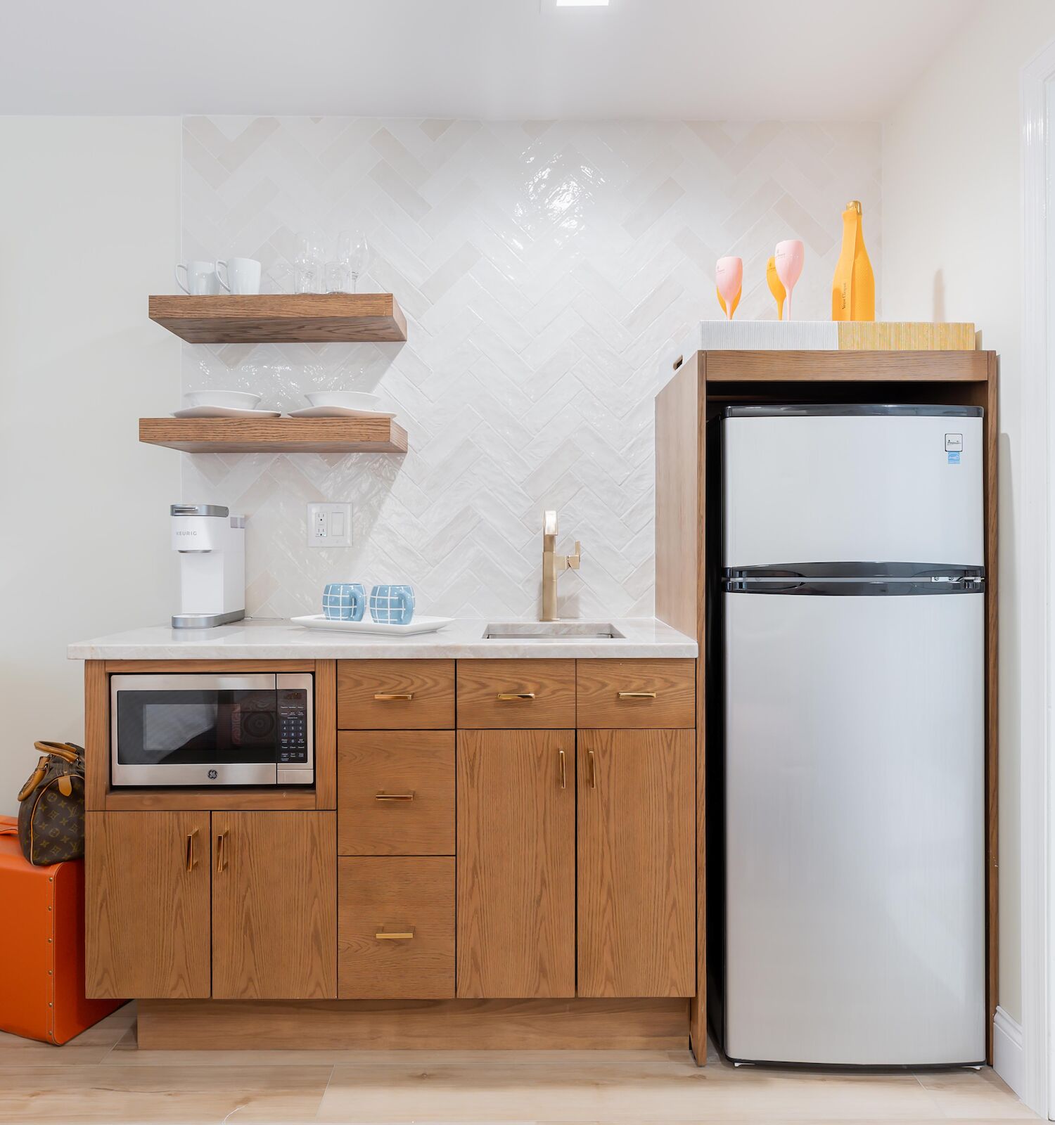 A modern kitchenette with a microwave, sink, fridge, open shelves, and orange accents, including a stool and decorative bottles.