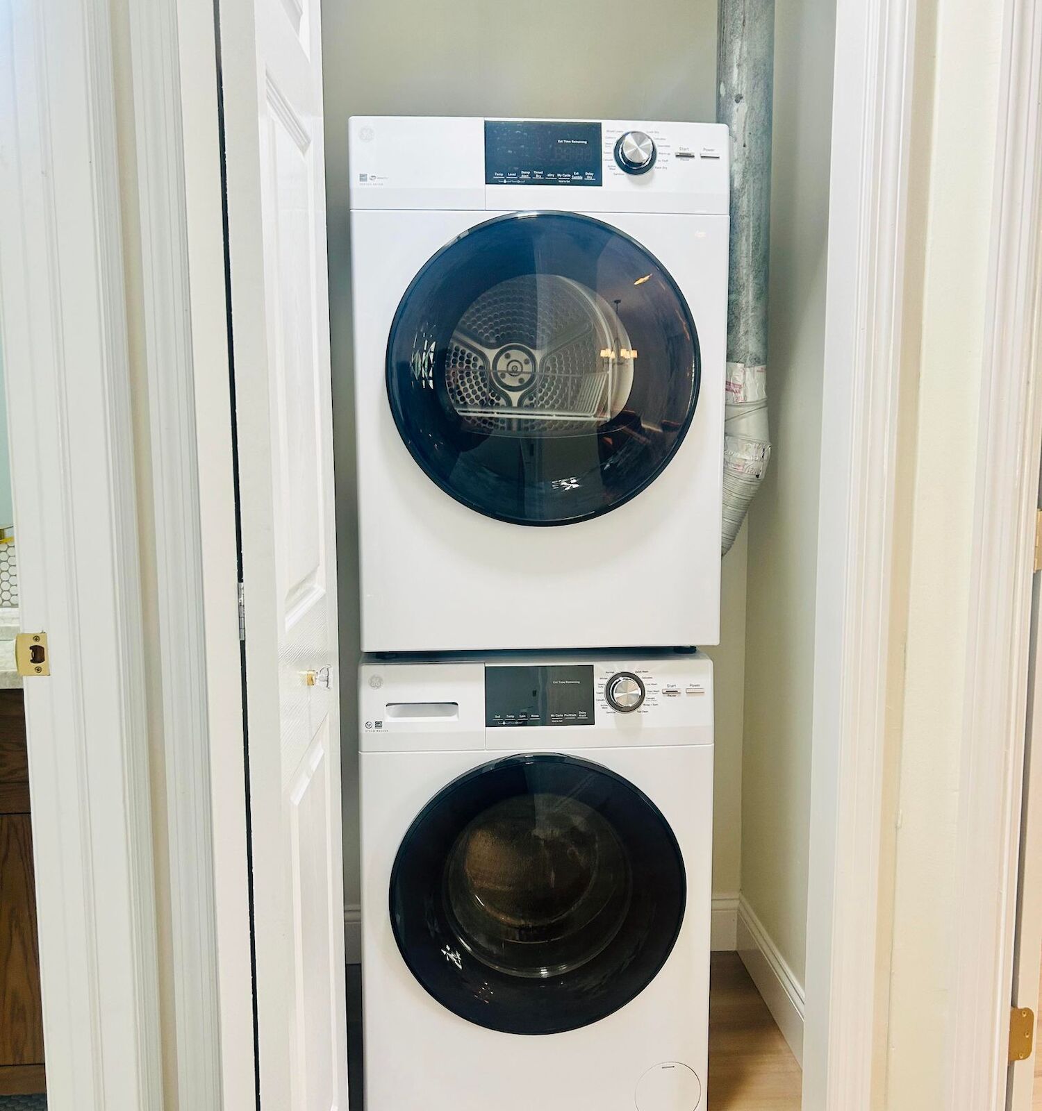 The image shows a stacked washer and dryer set in a small laundry closet with white folding doors and light-colored walls.