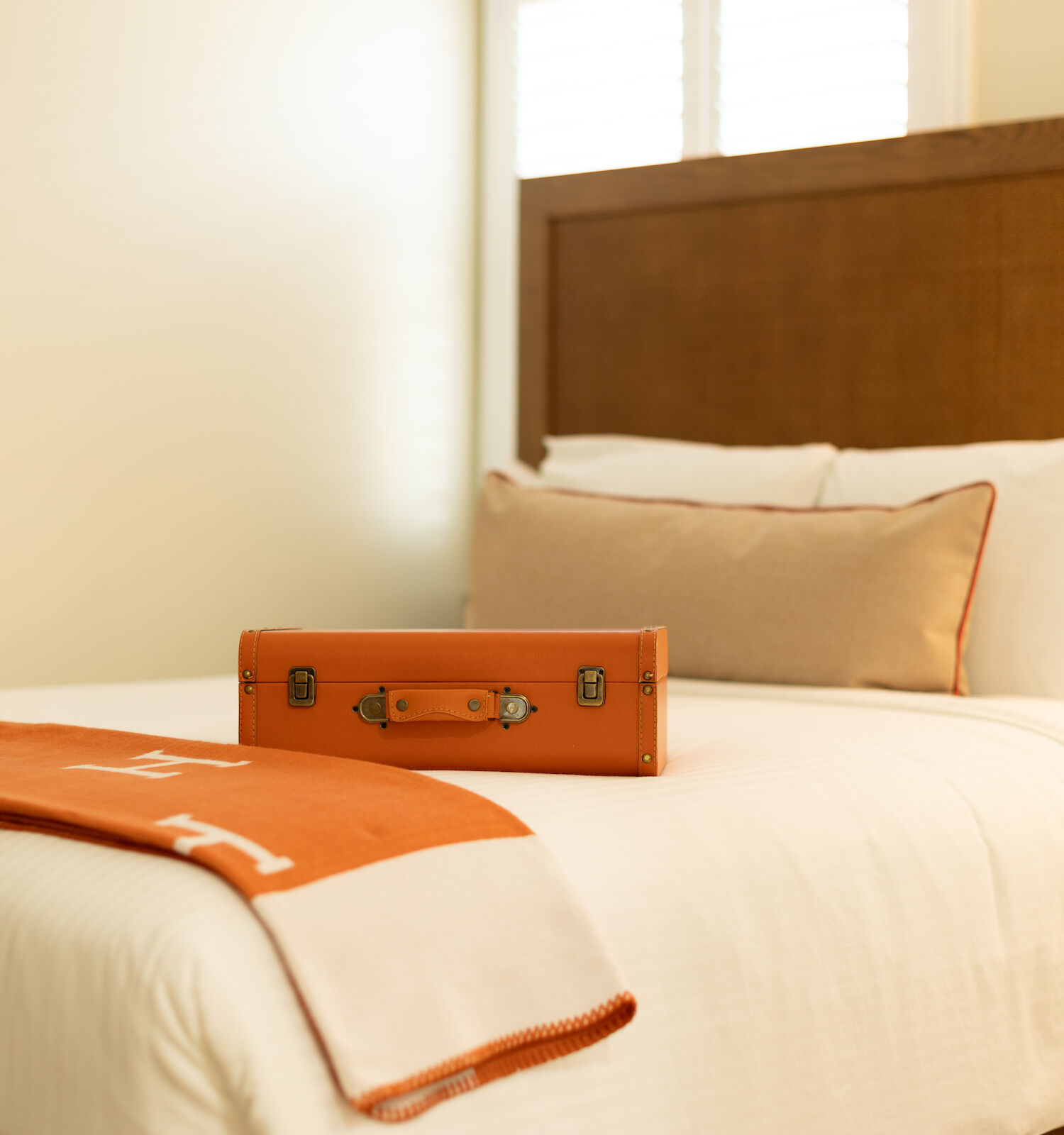 A cozy bed with white linens, an orange and white blanket, and a small orange suitcase placed on top.