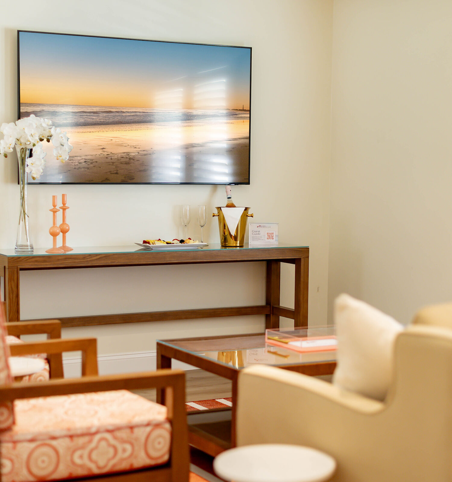 A cozy living room with a wall-mounted TV displaying a beach scene, comfortable chairs, a couch, table, and a console with decor items.