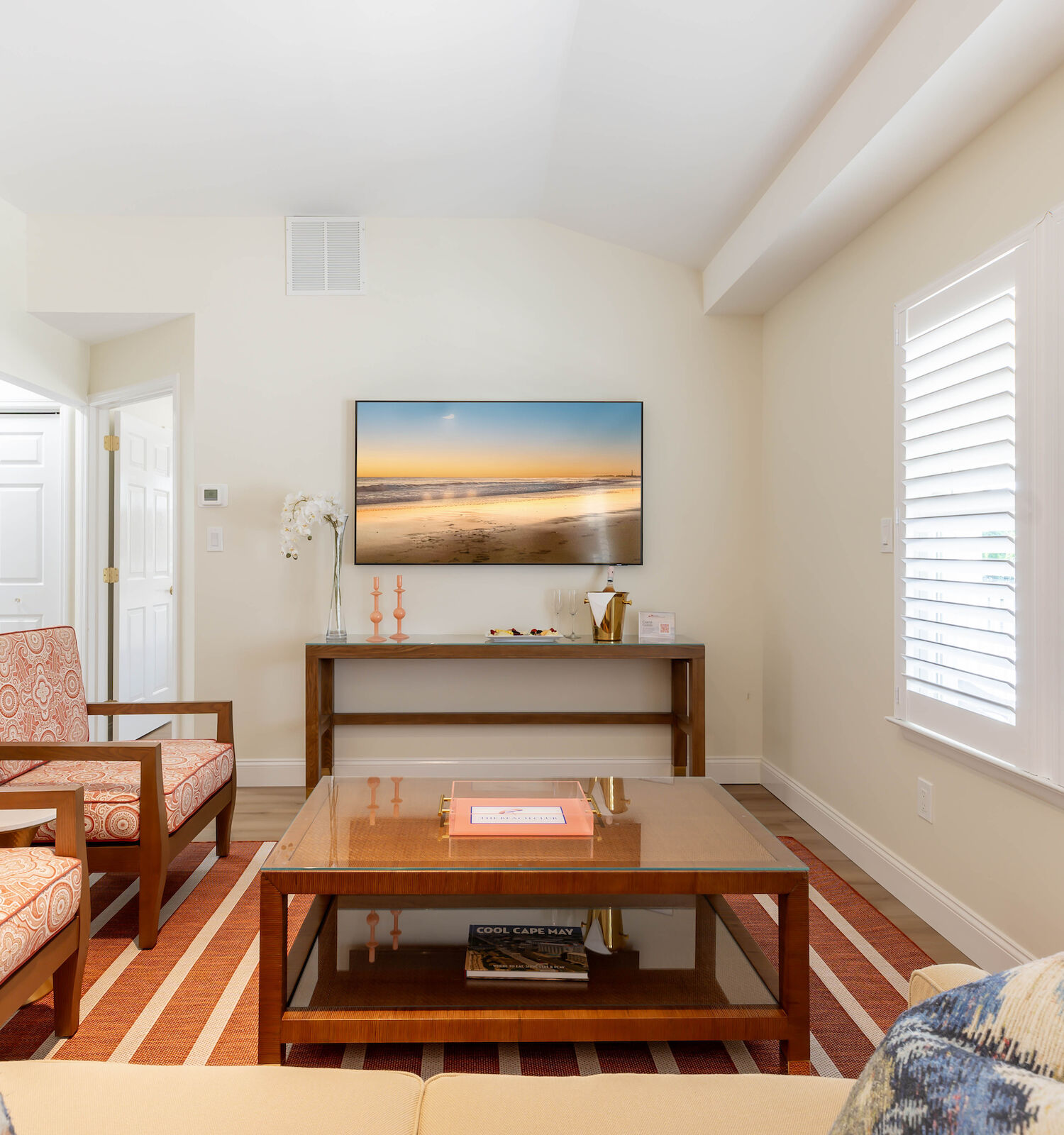 A cozy living room with beige walls, orange-patterned chairs, a wooden coffee table, and a wall-mounted TV displaying a beach scene.