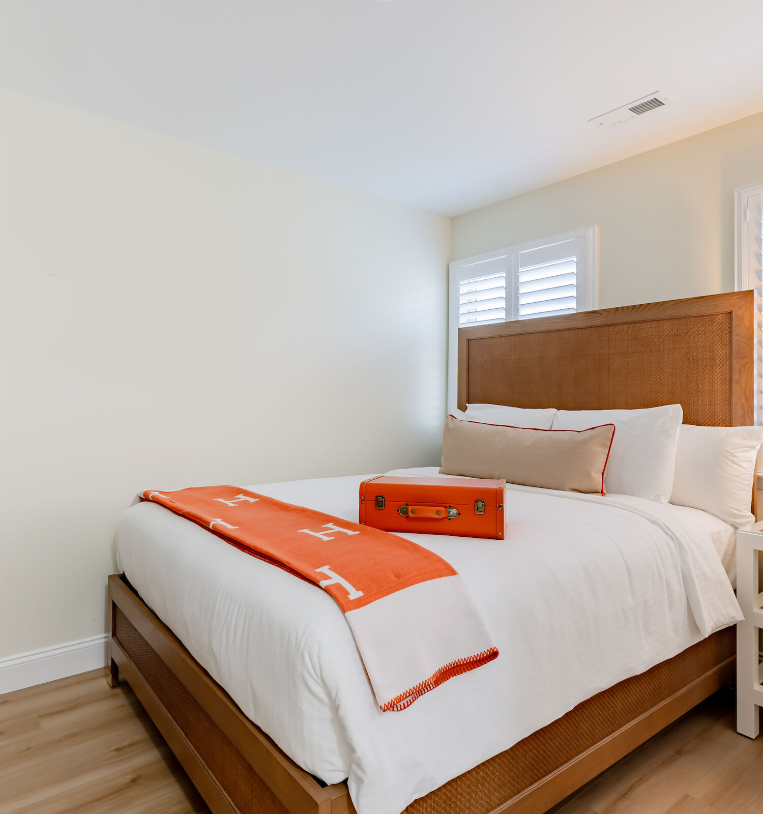 A bright bedroom with a large bed, wooden headboard, orange and white blanket, and suitcase; nightstand with lamp; dresser with bag.