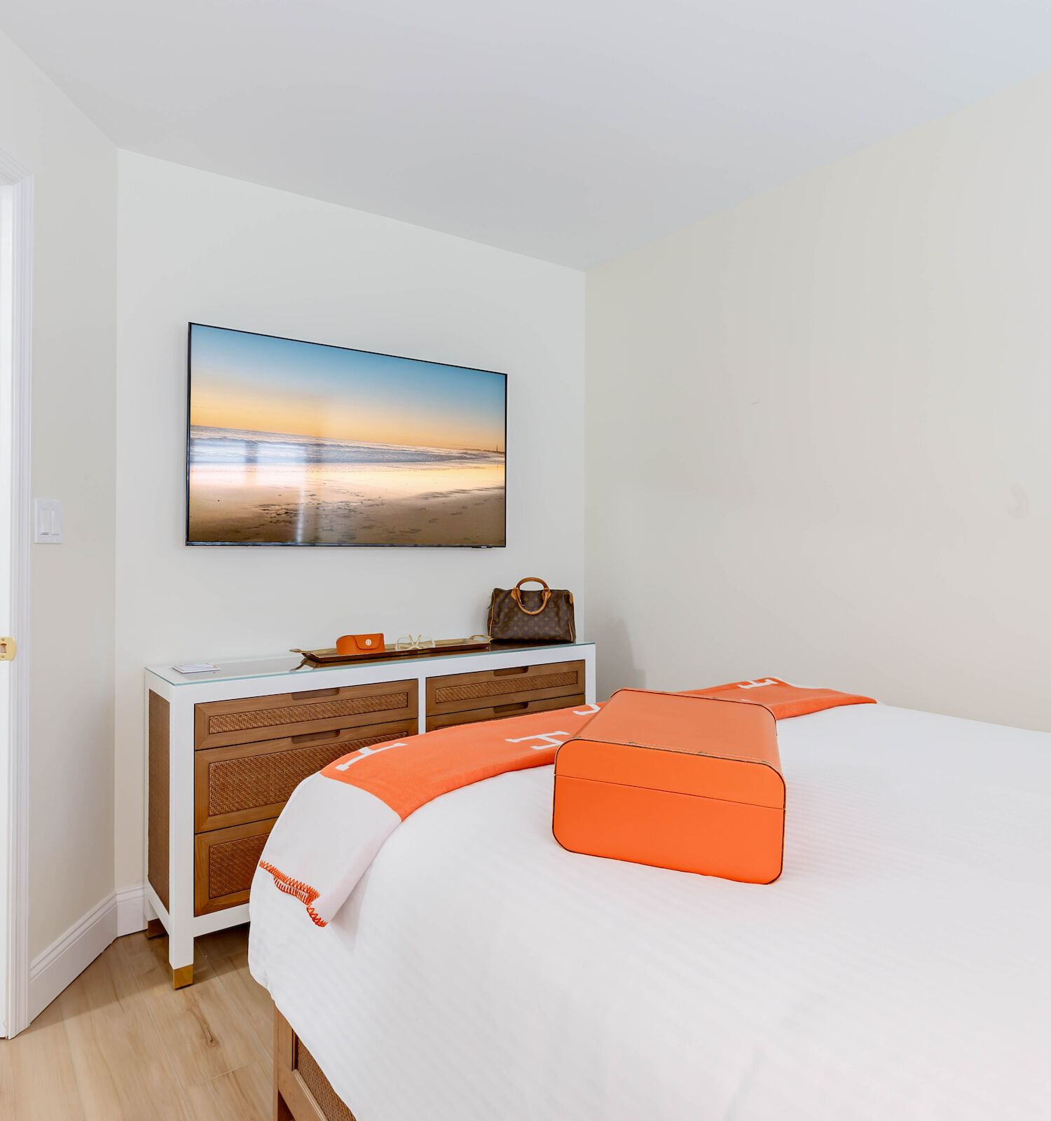 A modern bedroom with a neatly made bed, accented with orange pillows and blankets. There’s a sideboard, a mounted TV, and a hallway with décor.