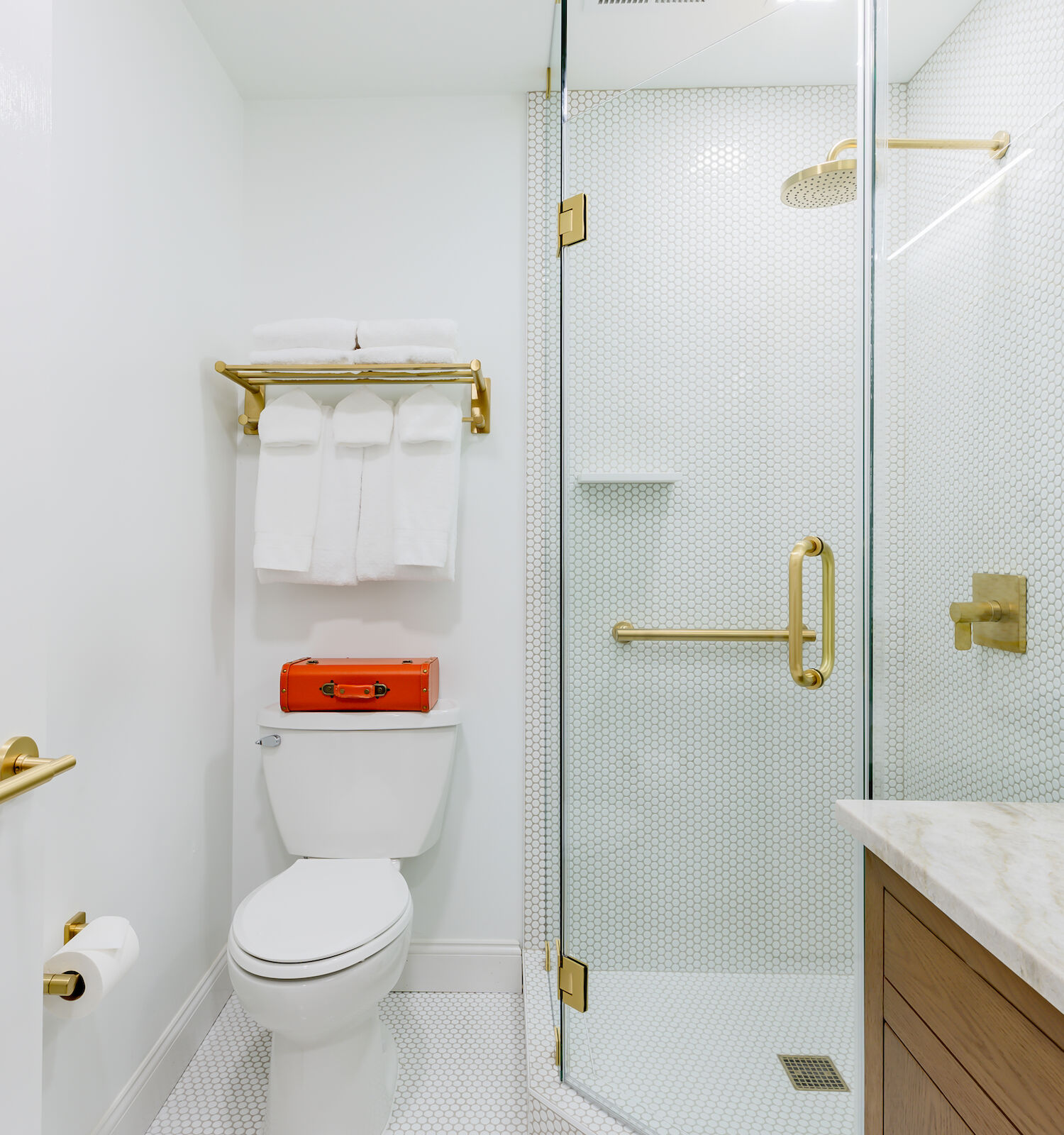 A modern bathroom with a glass shower enclosure, gold fixtures, a toilet with towels above, a sink with a countertop, and a roll of toilet paper.