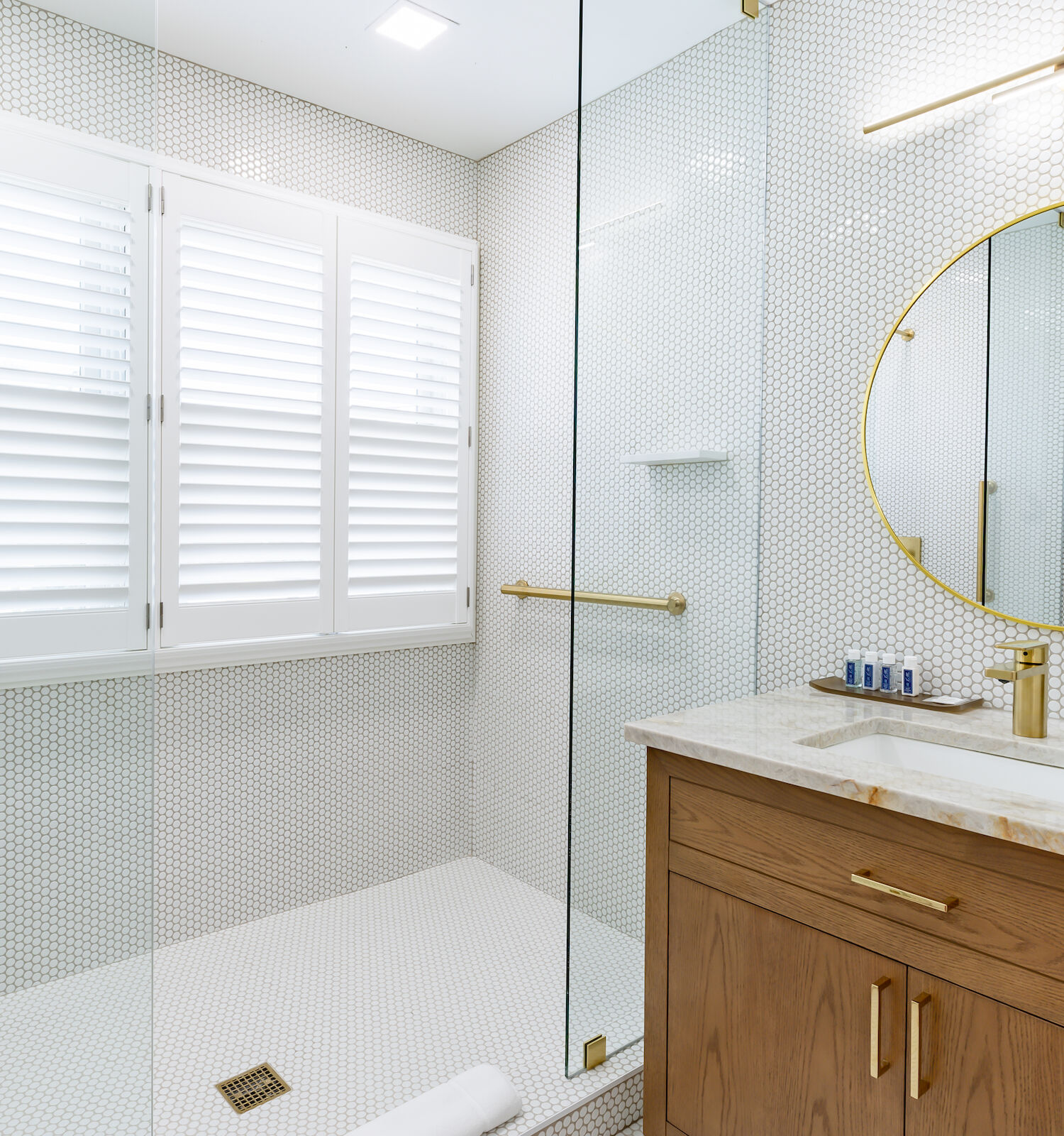 This image shows a modern bathroom with a glass shower, wood vanity, round mirror, gold fixtures, and white walls with a small tile pattern.