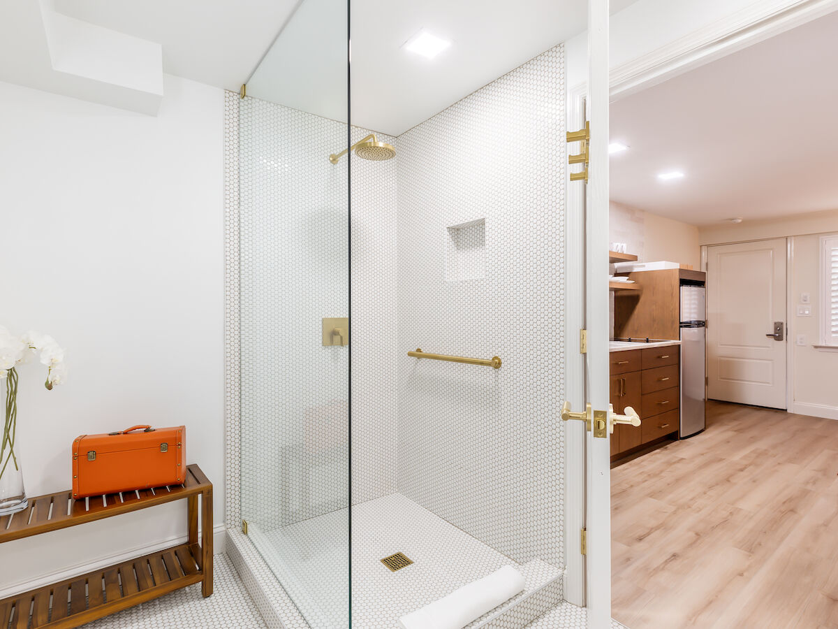The image features a modern bathroom with a glass shower, a wooden bench, a suitcase, and flowers. Visible adjoining room includes a kitchen.