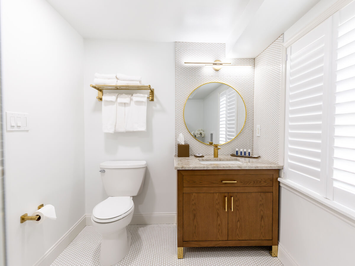 A modern, white bathroom with a wooden vanity, round mirror, toilet, gold fixtures, and folded towels on a shelf, with natural light from shutters.
