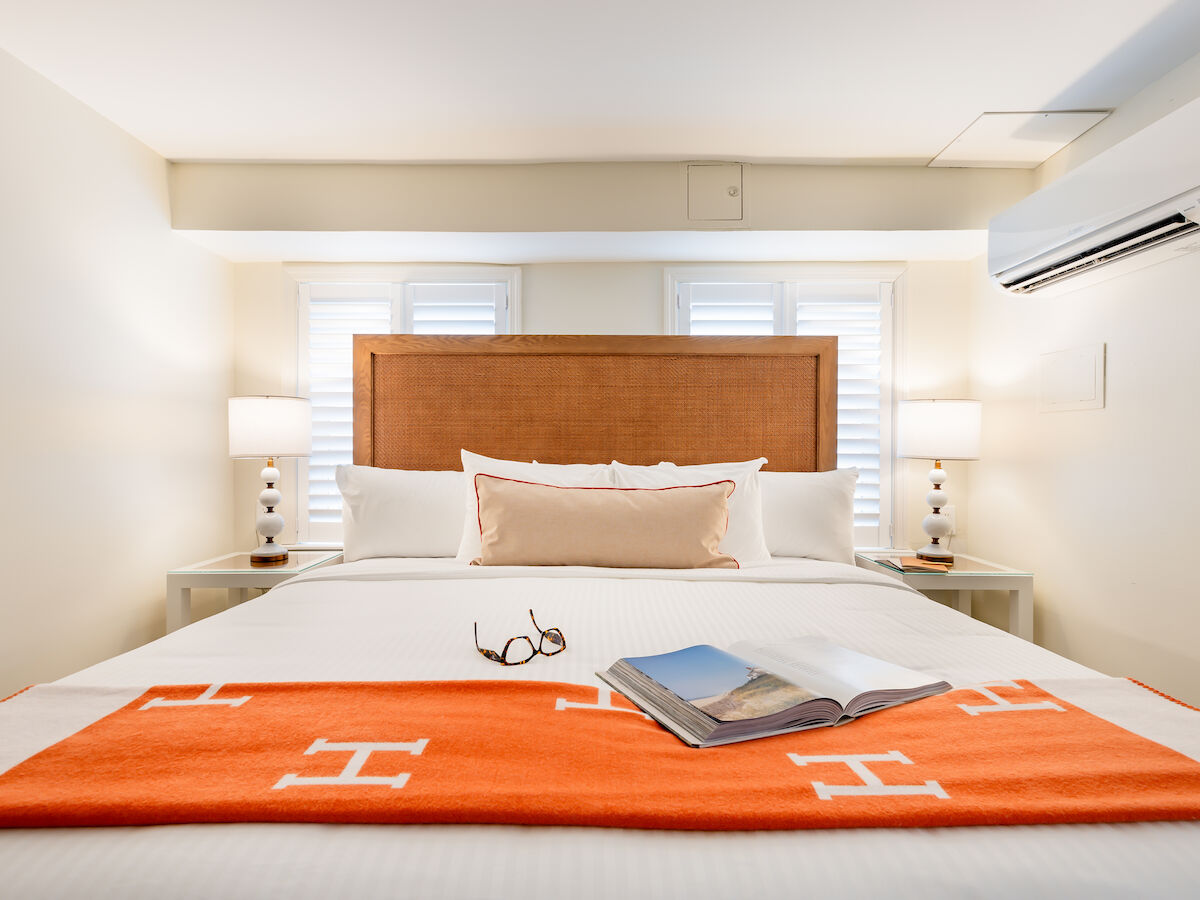 A neatly made bed with an orange blanket, pillows, a book, and eyeglasses. Two bedside lamps and an air conditioner are visible.