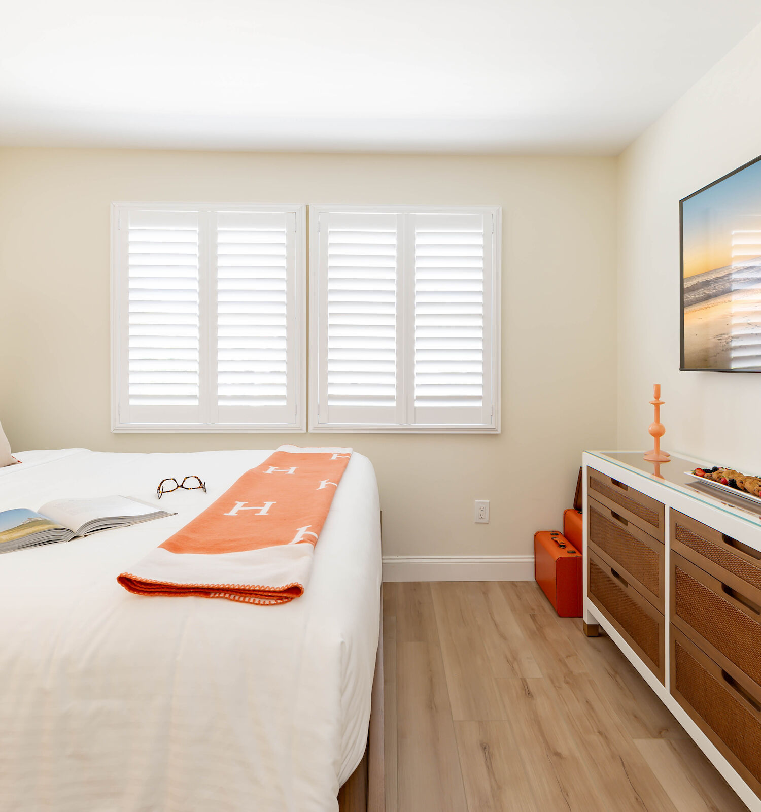 A minimalist bedroom features a bed with white linens and orange accents, a TV on the wall, and a dresser with decor items including a wine glass and bottle.