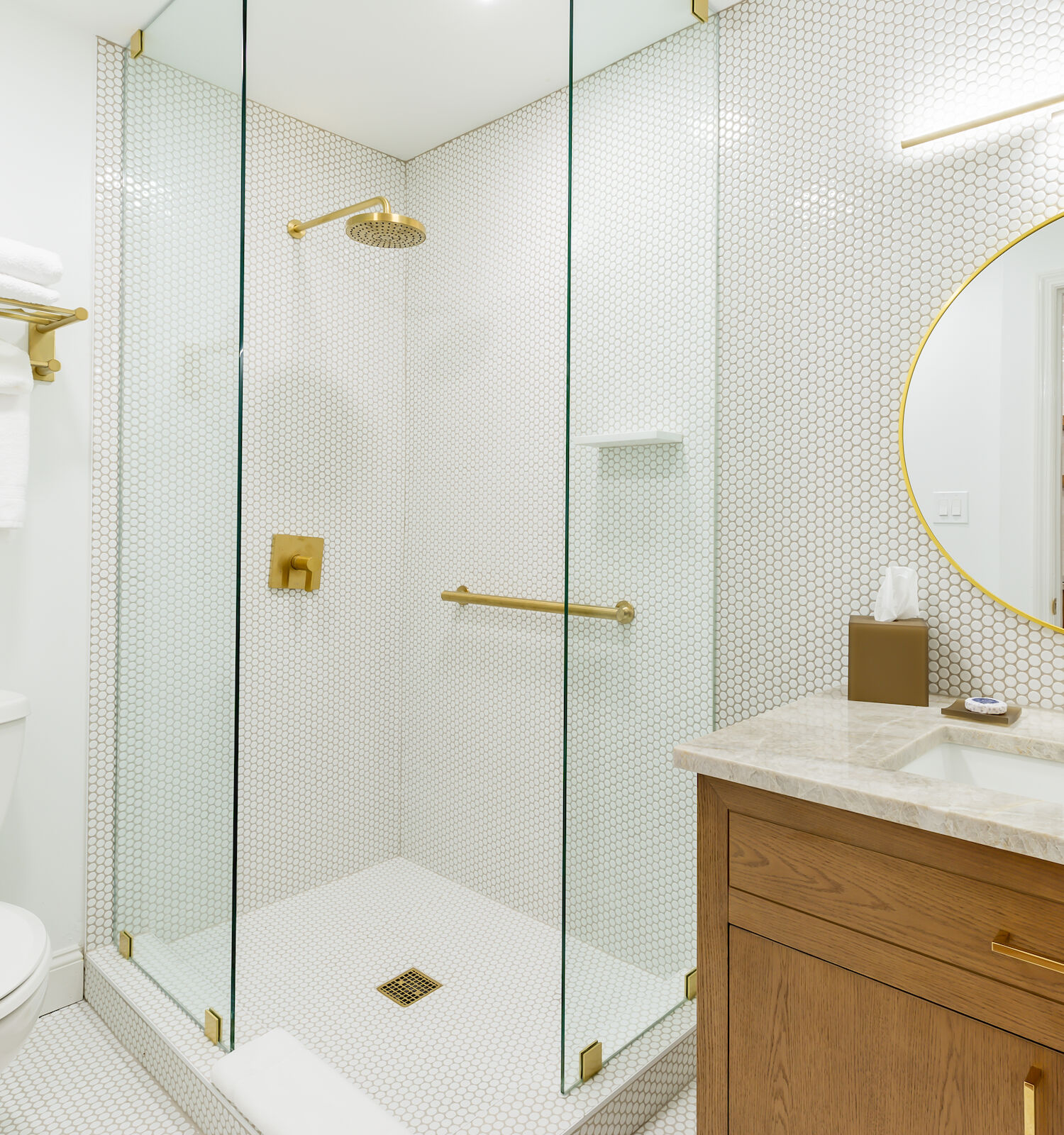 A modern bathroom with a glass shower, gold accents, a round mirror, a toilet, and a wooden vanity with a light countertop.
