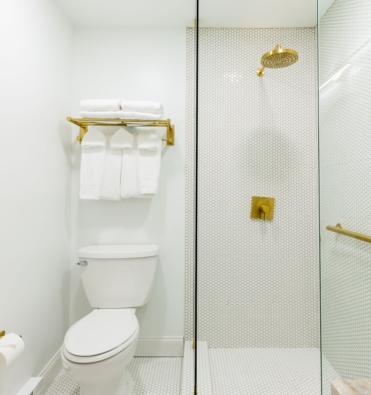 A modern bathroom with white wall tiles, a toilet, gold shower fixtures, a glass-enclosed shower, and a towel rack with white towels.