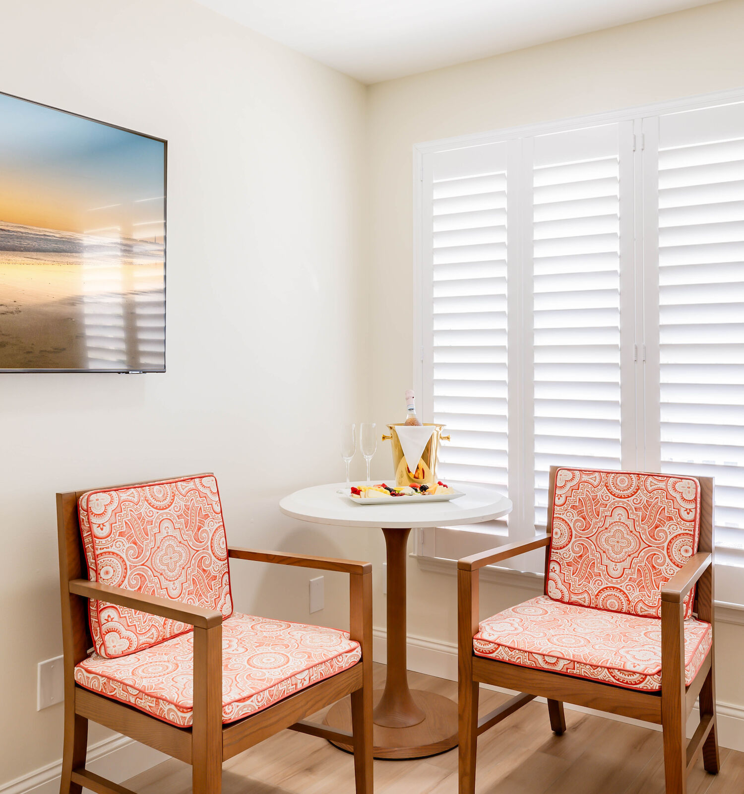 A small, bright room with two patterned chairs around a round table, a tray with a coffee pot and cups, a beach photograph on the wall, and white shutters.