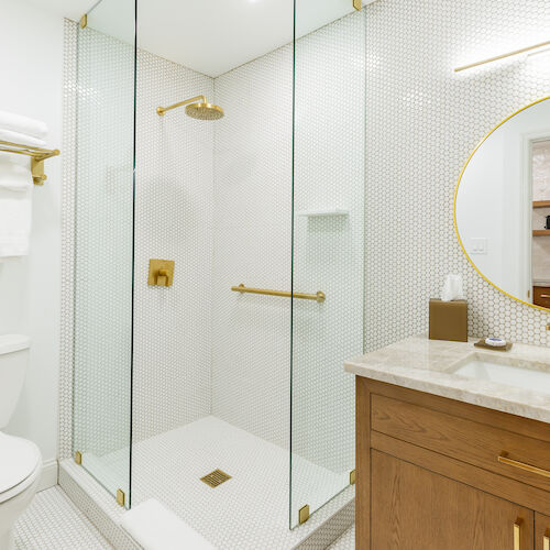 A modern bathroom with a glass shower, gold fixtures, a round mirror, wooden vanity, and toilet. Towels are neatly hung on a rack.