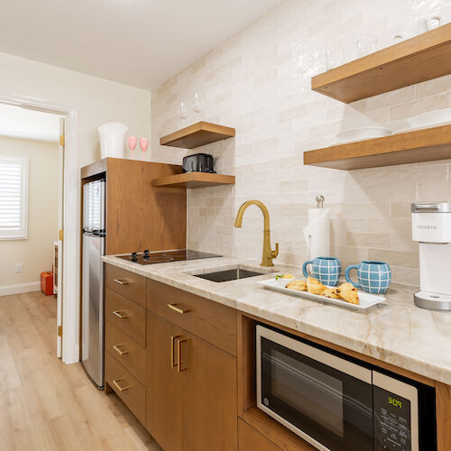A compact kitchen with wooden cabinets, a microwave, and shelves is adjacent to a bedroom through an open door.