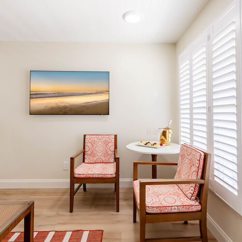 A small, cozy room with two patterned chairs, a small table set for snacks, and a beach photo on the wall. The room has shutters on the windows.