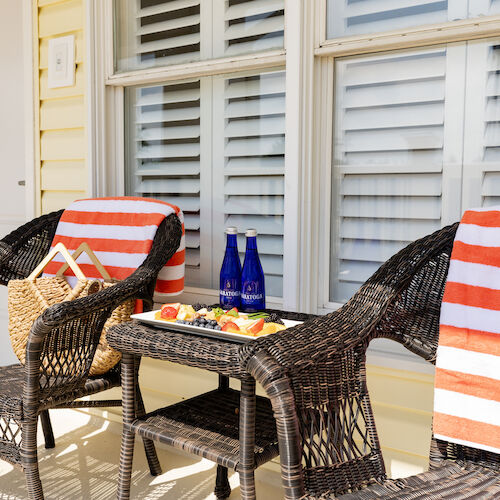 Two wicker chairs on a porch with striped towels, a table with snacks, blue bottles, a hat, and a woven bag with books, framed by window shutters.
