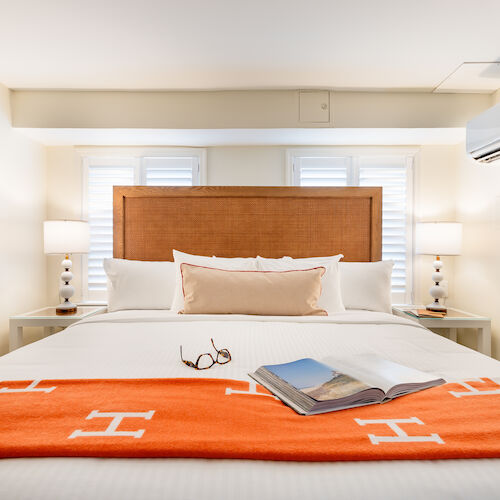 A neatly made bed with an orange throw blanket, an open book, and glasses on top, flanked by two bedside tables and lamps in a well-lit room.
