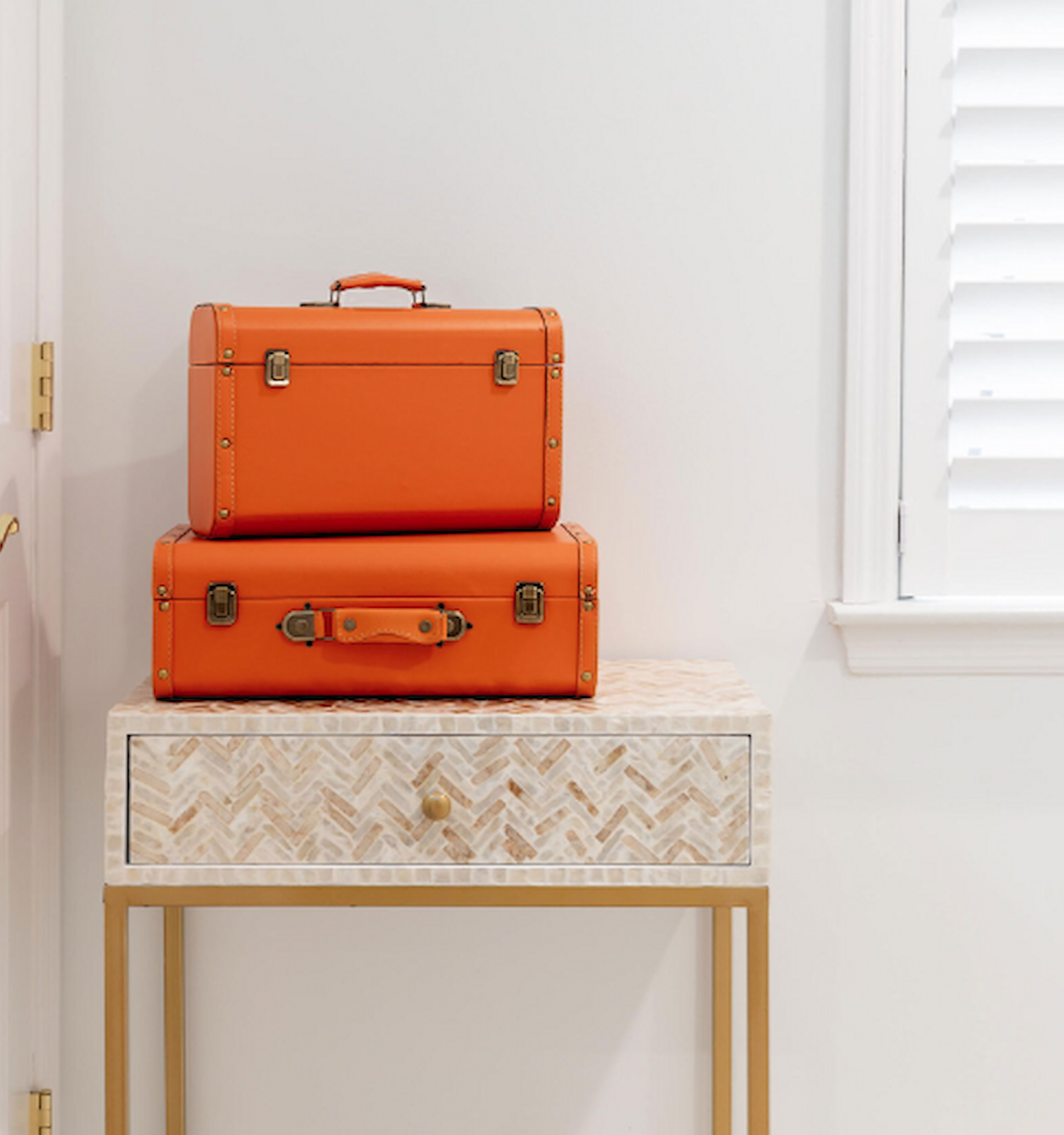 Two orange suitcases are stacked on a small table with a herringbone pattern in a minimalist room with white walls and a shuttered window ending the sentence.