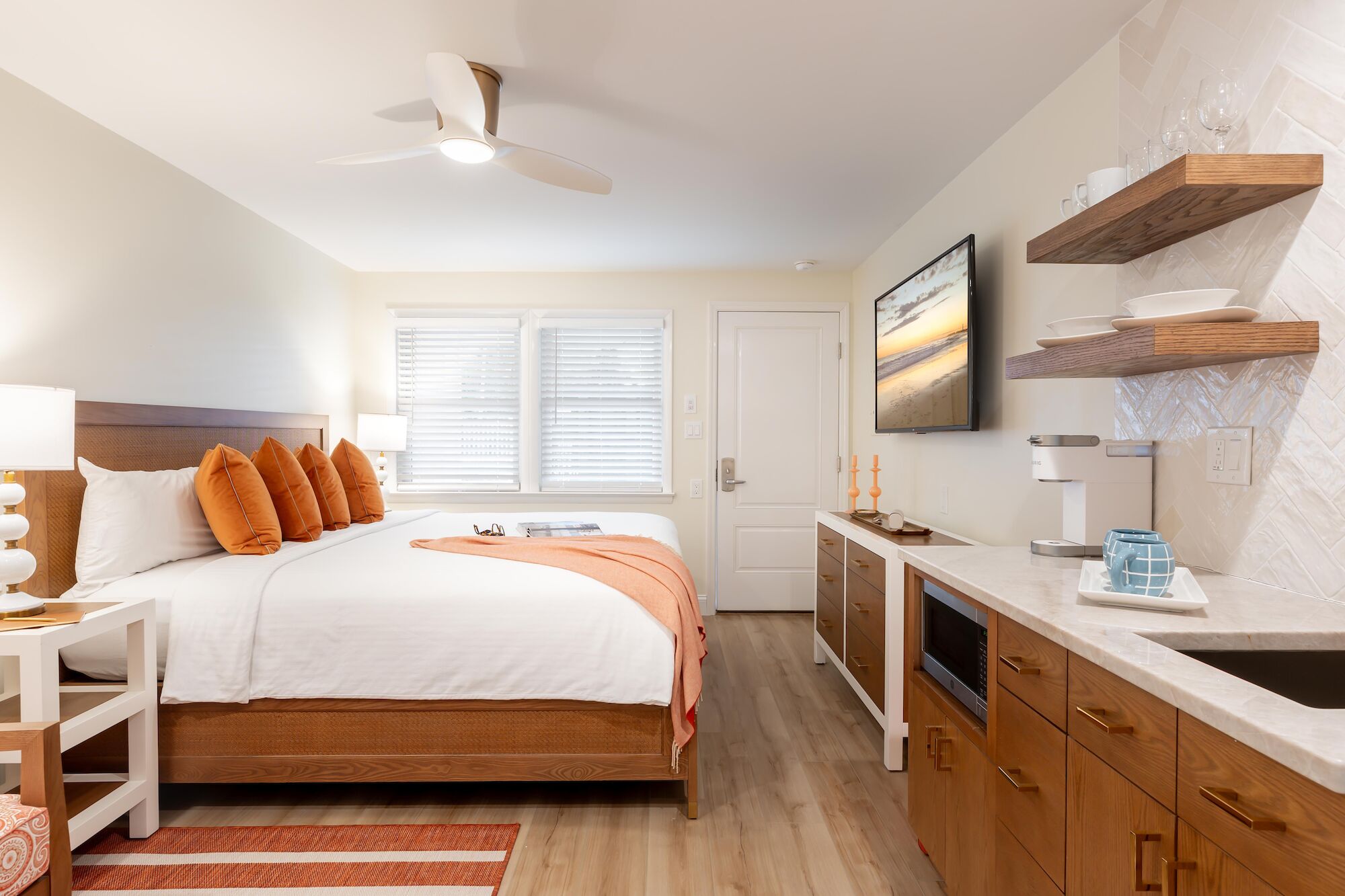 A modern, cozy bedroom with a large bed, orange accents, wall-mounted TV, ceiling fan, and a compact kitchenette area.