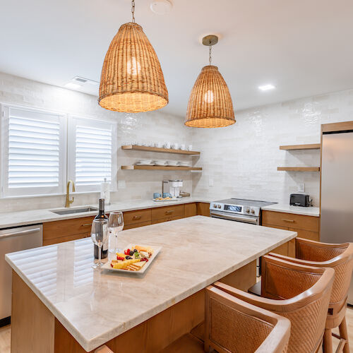 A modern kitchen with a central island, wicker chairs, hanging pendant lights, stainless steel appliances, and a platter of food on the countertop.