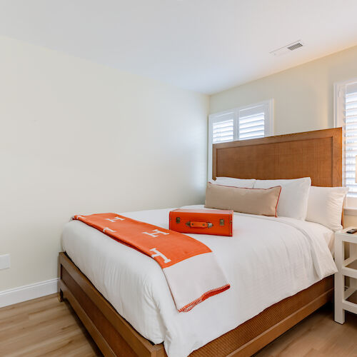 A bedroom with a neatly made bed, an orange throw and pillow, a lamp on a bedside table, and a dresser with a bag.