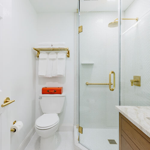 A bright, modern bathroom featuring a toilet, a glass-enclosed shower with gold fixtures, a towel rack with folded towels, and a vanity with a marble countertop.
