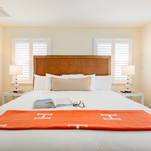 A neatly made bed with white linens, an orange blanket at the foot, a book and glasses placed on the bed, flanked by two lamps on side tables.