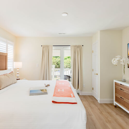 A bright bedroom features a large bed, a dresser with a TV, glass doors leading to a balcony, and a serene decor with white and wooden tones.