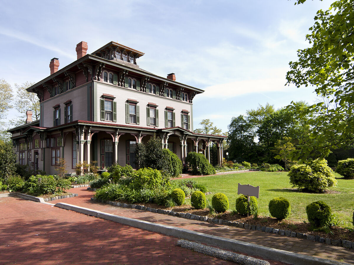 The image shows a large, historic house with a manicured lawn, diverse greenery, and a long driveway extending from the foreground.