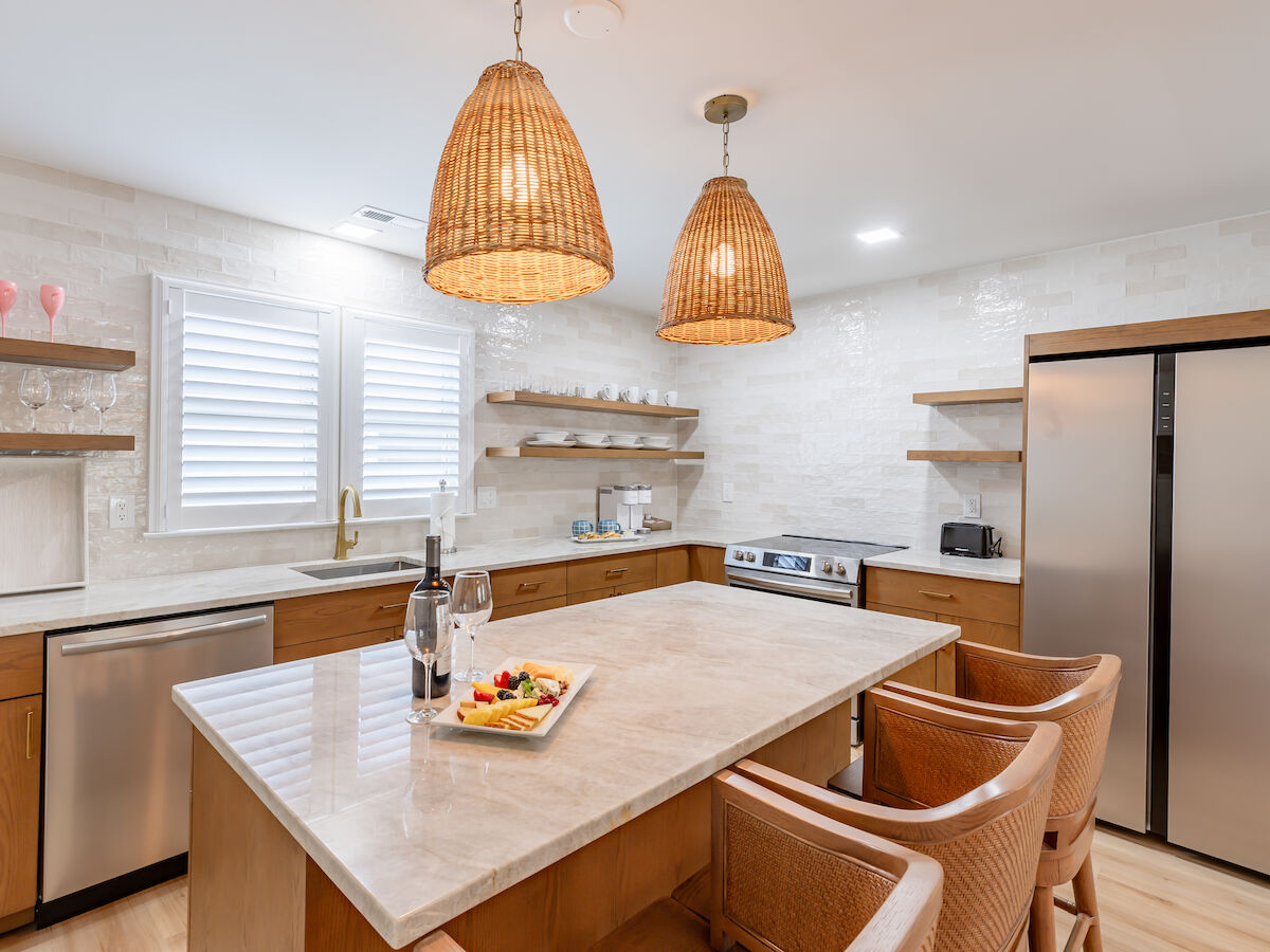 A modern kitchen with an island, wicker chairs, pendant lights, double-door fridge, dishwasher, oven, wine glasses, and a fruit tray on the countertop.