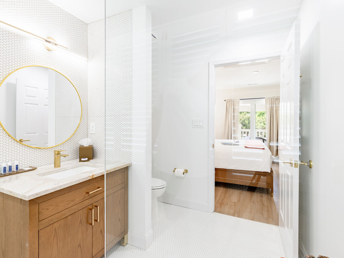 The image shows a modern bathroom with a wooden vanity, round mirror, and gold fixtures, leading to a bright bedroom with a bed and large windows.