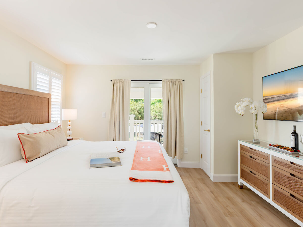 A bright bedroom with a large bed, TV, dresser, and French doors leading to a balcony with a view. The decor is modern and minimalist.