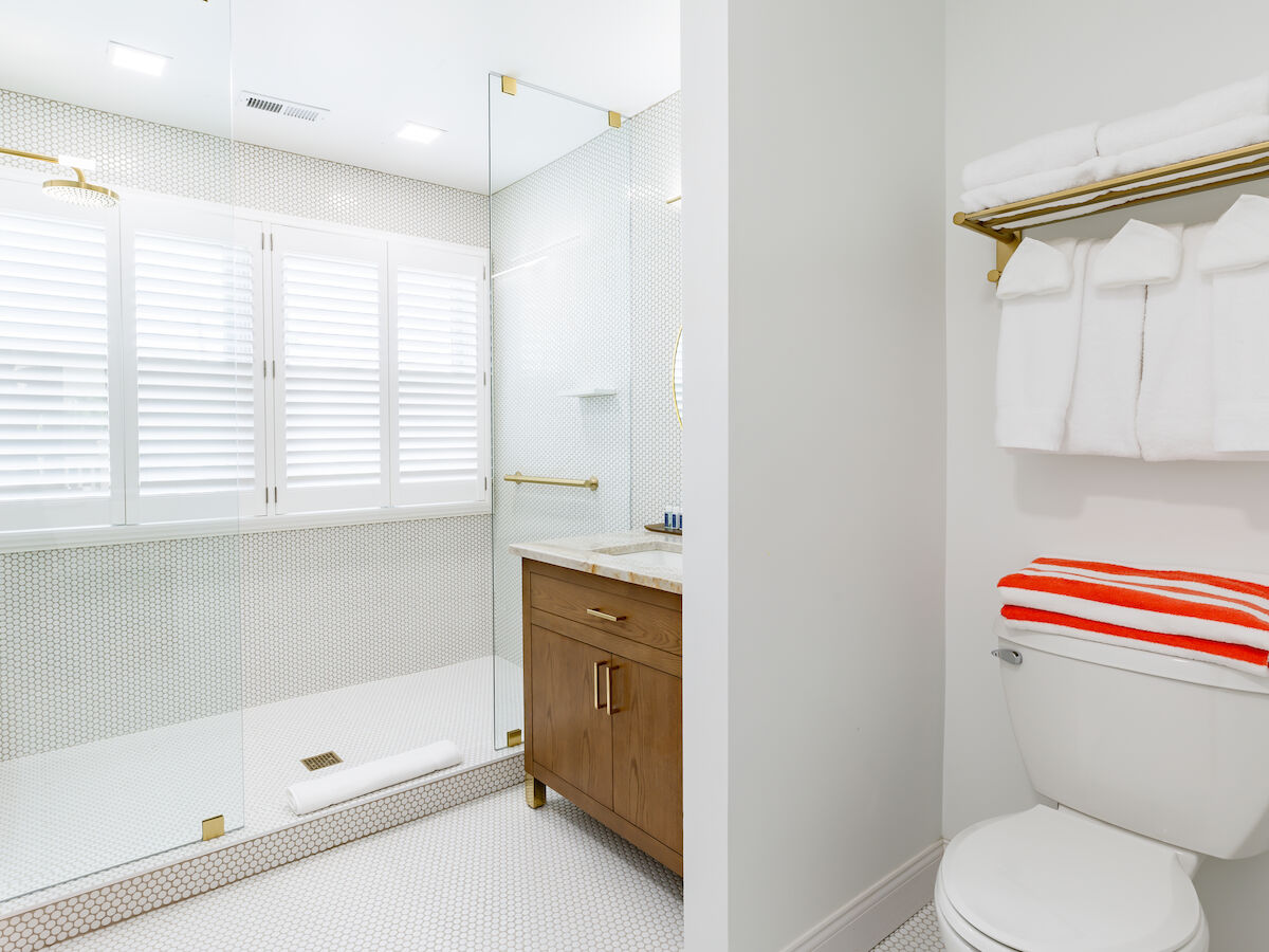 A modern bathroom with a walk-in shower, vanity, toilet, towels, and window shutters. Red and white striped towel adds a pop of color.