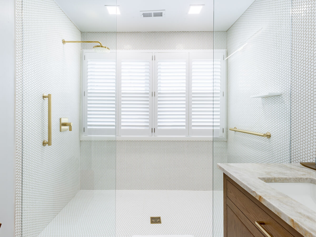 A modern bathroom with a glass-enclosed shower, gold fixtures, white tiles, a vanity with a marble countertop, and a round mirror.