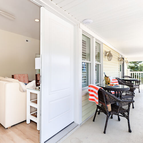 A cozy porch with wicker chairs and striped towels overlooks a scenic view; an open door reveals a plush living room inside.