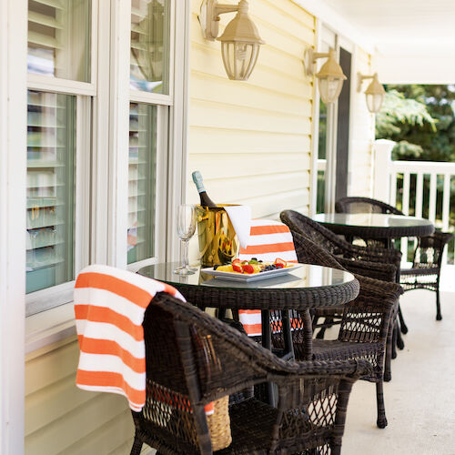 A cozy porch with wicker chairs, orange-striped towels, a table set with wine and bananas, and wall-mounted lanterns beside yellow siding.