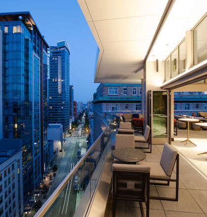 A modern balcony with seating overlooks a cityscape filled with illuminated buildings and busy streets at dusk.