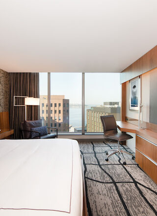 A modern hotel room with a large bed, a wall-mounted TV, a work desk with a chair, and a window offering a city view.