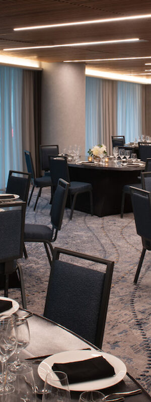 The image shows an elegantly set dining room with round tables, black chairs, and detailed table settings, including glasses, napkins, and centerpieces.