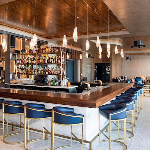 The image shows a modern bar with blue stools and a stocked bar counter. Pendant lights hang from the ceiling, with a cozy seating area nearby.