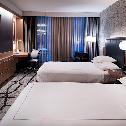 A modern hotel room with two neatly made beds, a wall-mounted TV, a work desk, and stylish lighting.
