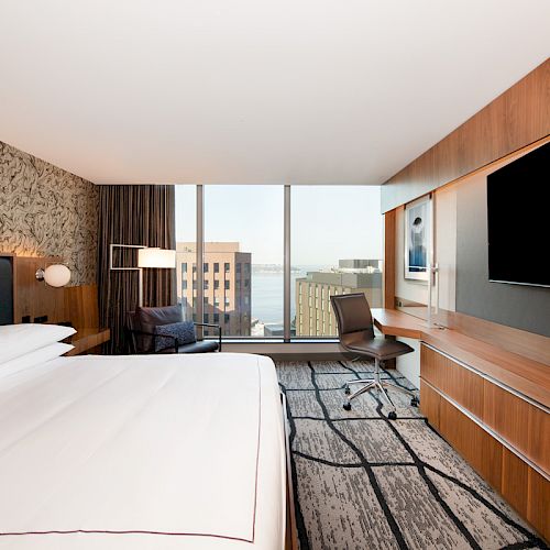 A modern hotel room with a large bed, wall-mounted TV, desk, chair, window with a city view, and contemporary decor.